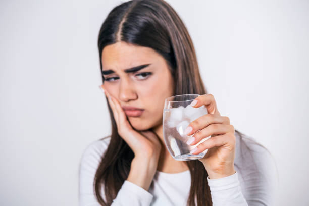 Young woman with sensitive teeth and hand holding glass of cold water with ice. Healthcare concept. woman drinking cold drink, glass full of ice cubes and feels toothache, pain